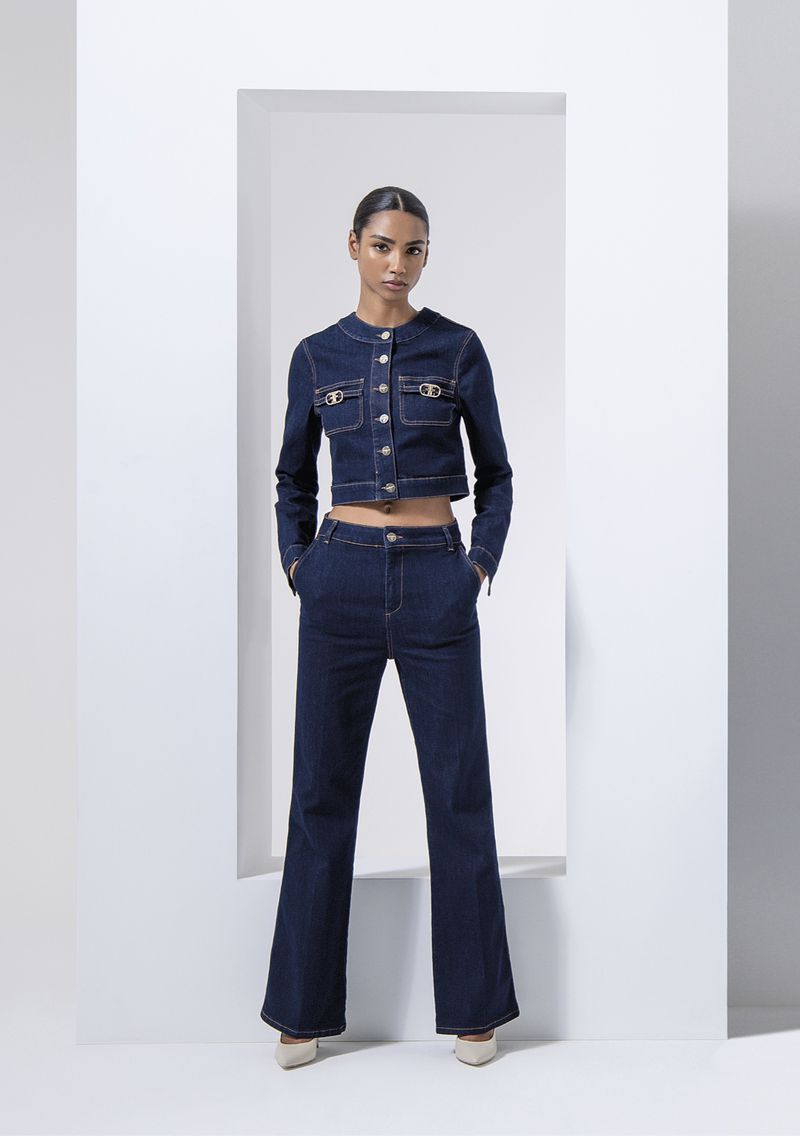 Fashion model posing in a denim jacket and bell-bottom jeans against a white background with architectural structure.
