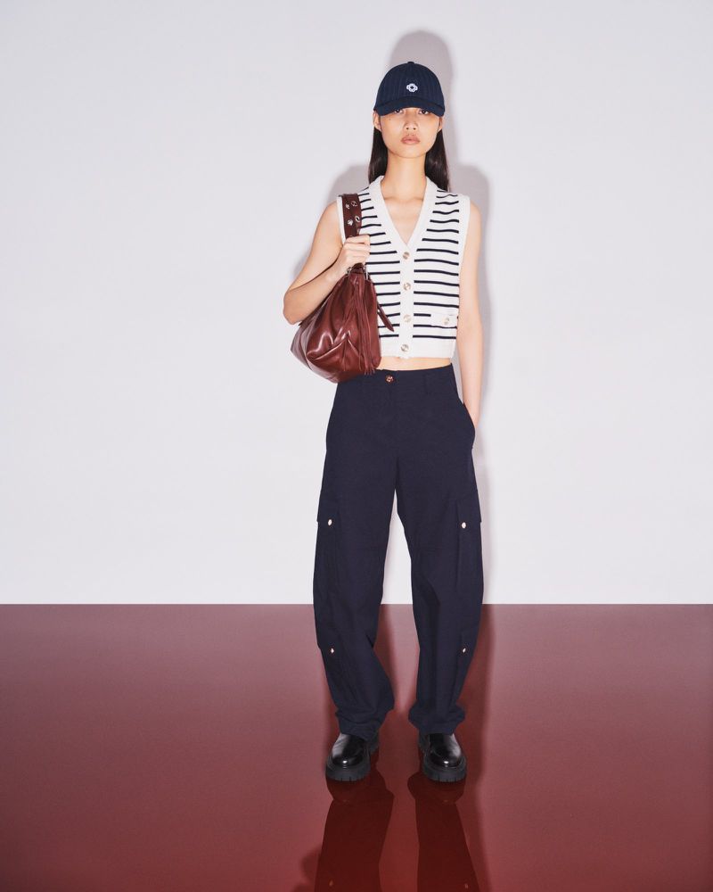 Young woman in a sleeveless sweater vest, navy pants, and baseball cap poses with a shoulder bag against a gradient studio backdrop.