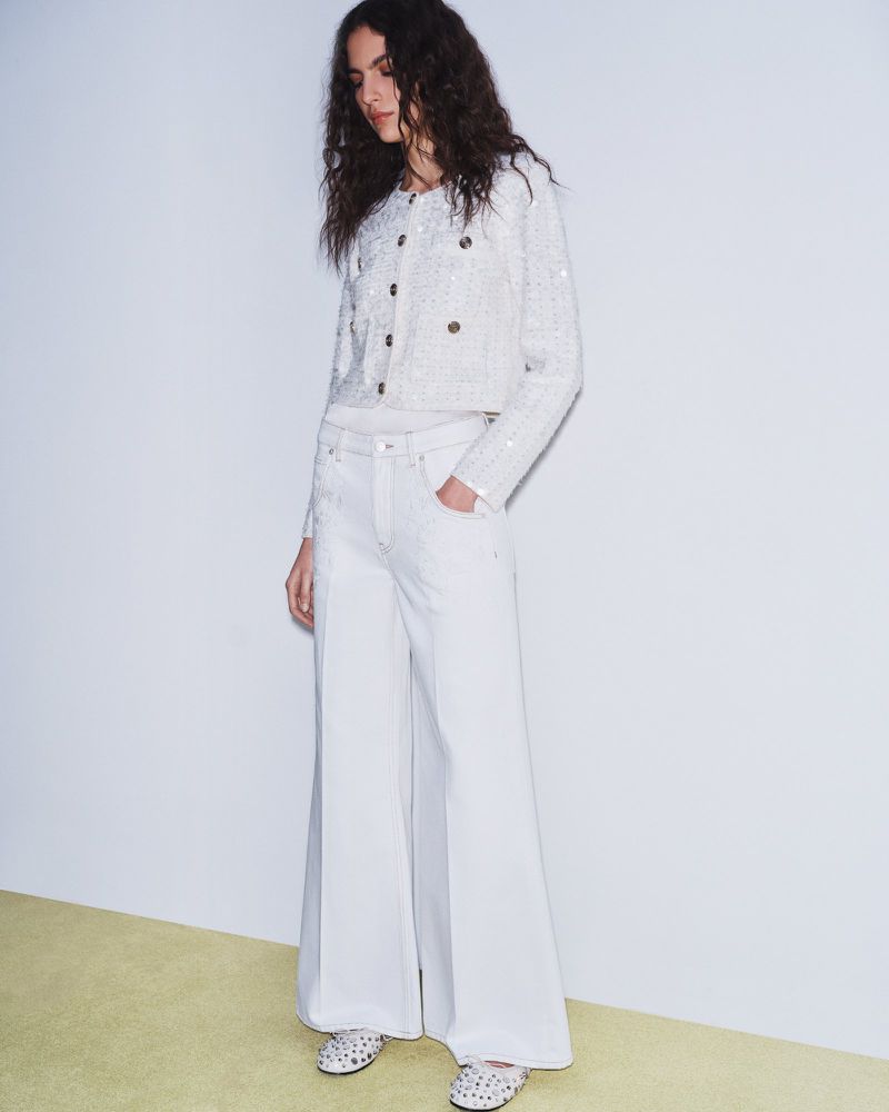 A woman in a white sequined jacket and wide-leg jeans stands against a white wall in a fashion shot emphasizing style and design.