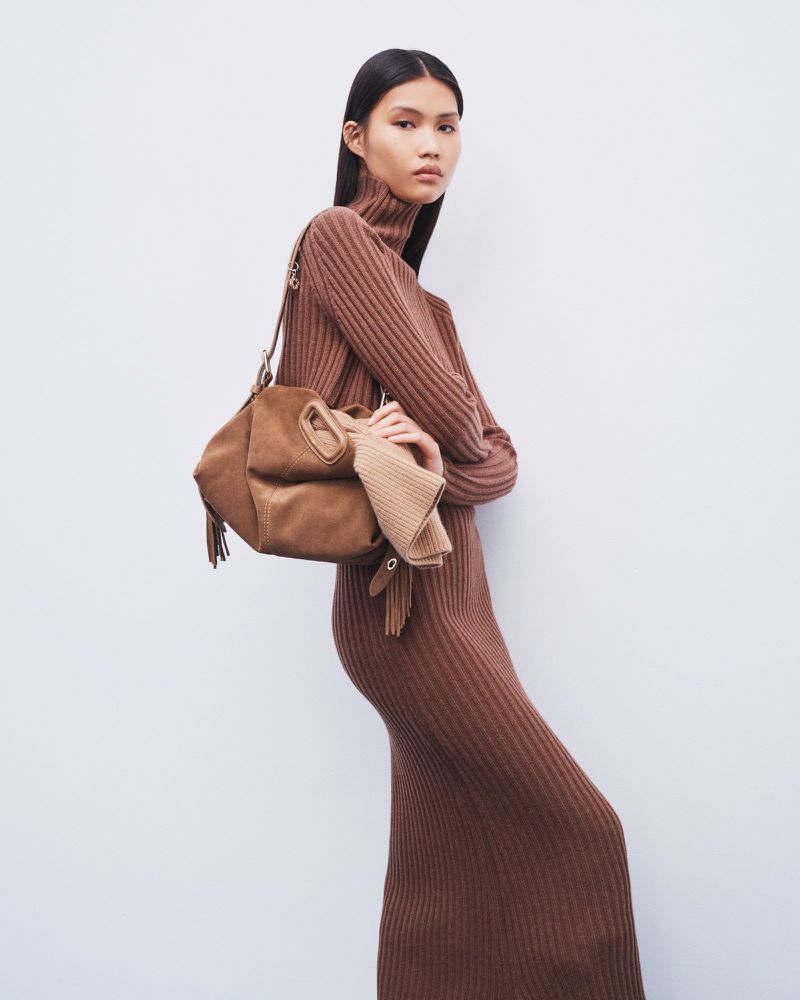 Fashionable woman in a brown knit dress poses in front of a white wall, carrying a tan suede handbag with fringe details.