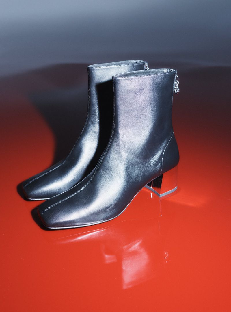 Studio shot of metallic silver ankle boots with square toes and clear block heels on a red reflective surface with a grey background.