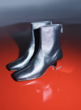Studio shot of metallic silver ankle boots with square toes and clear block heels on a red reflective surface with a grey background.