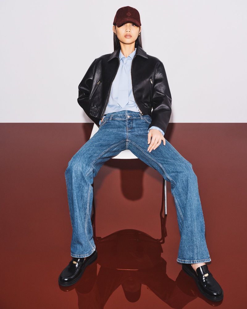 Woman sits in a photo studio wearing a baseball cap, cropped black leather jacket, blue jeans, and black patent leather loafers.
