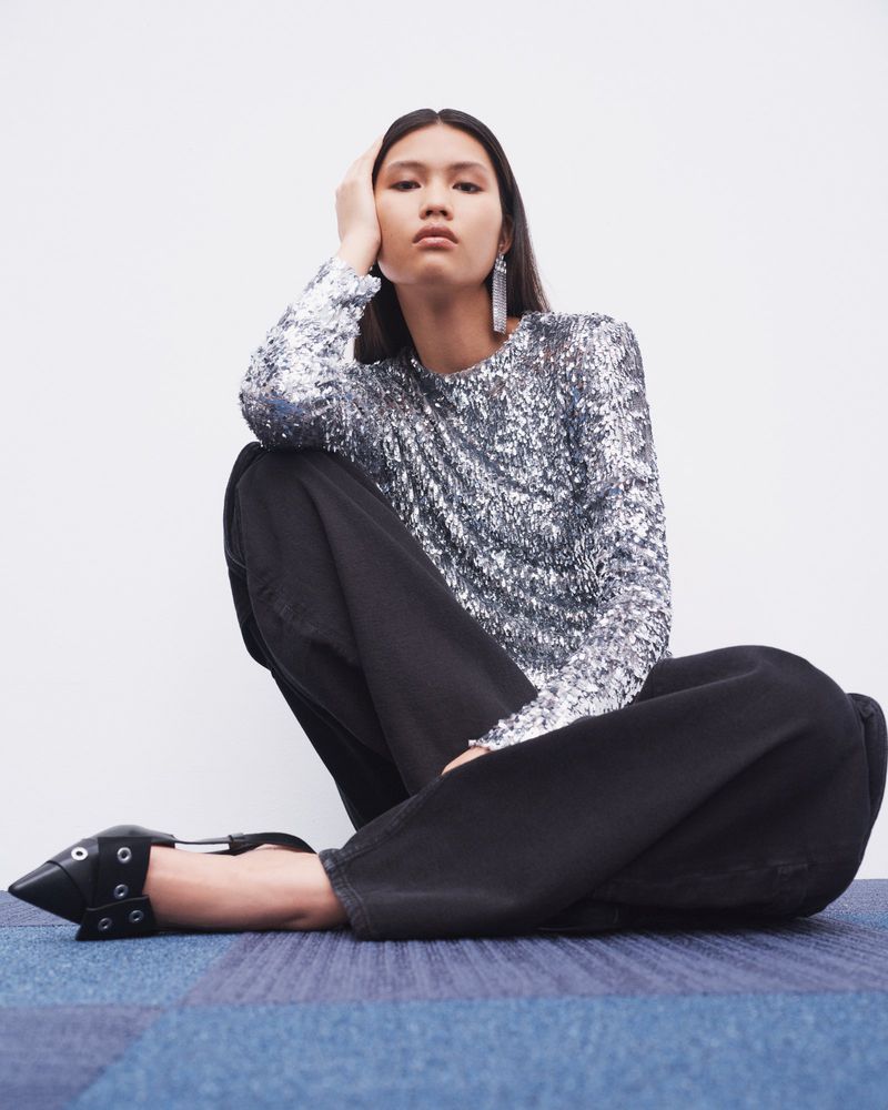 Fashionable Asian woman in a silver sequin top and black jeans sits casually against a white wall, showcasing modern style.
