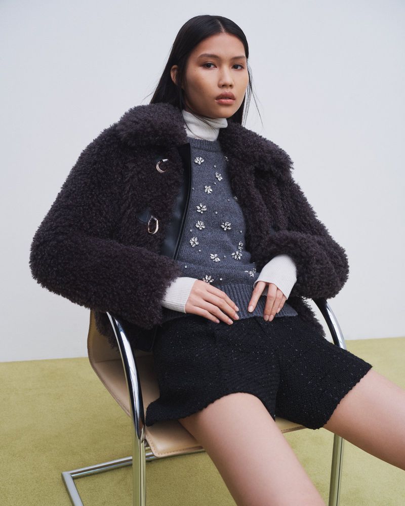 Jinrong Huang seated in a chrome chair wearing a dark fleece jacket, embellished grey sweater, and black shorts for Maje FW24.