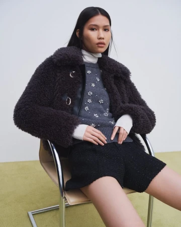 Jinrong Huang seated in a chrome chair wearing a dark fleece jacket, embellished grey sweater, and black shorts for Maje FW24.
