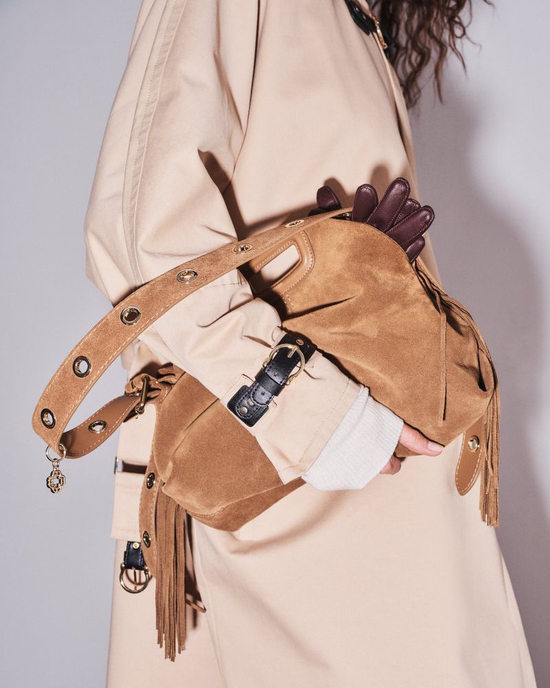 Close-up of a person wearing a beige trench coat holding a brown suede purse with tassels in a studio setting with soft, neutral lighting.