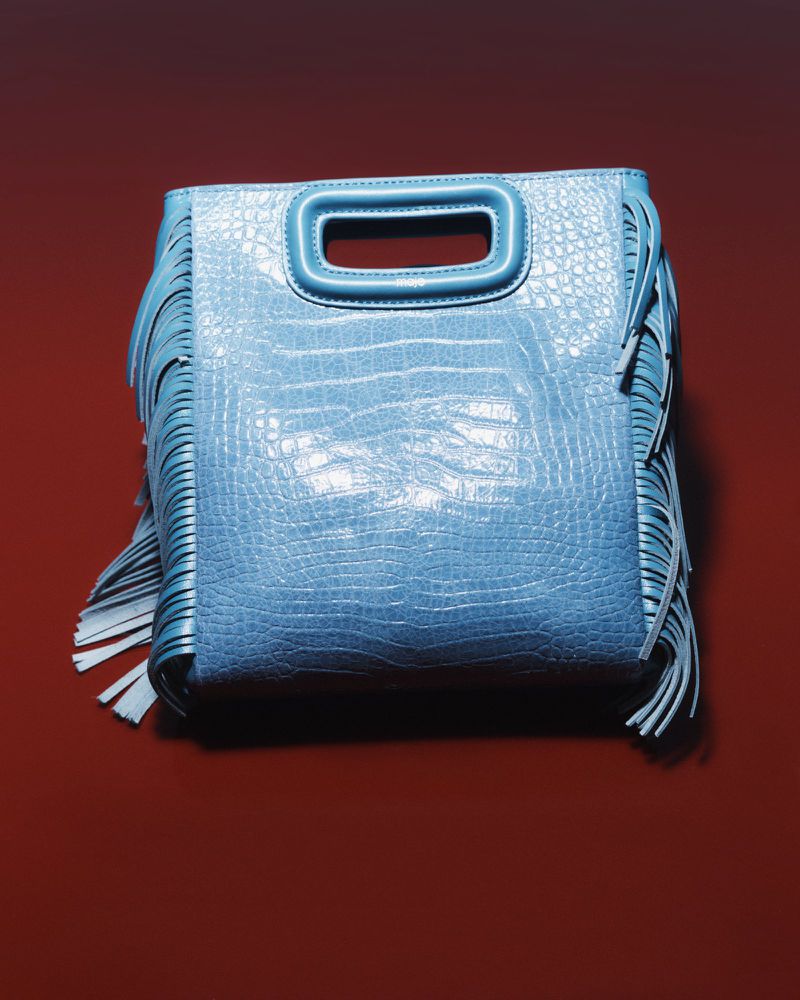 Fashionable blue alligator-print leather handbag with fringe detail against a dark red background in a studio product shot.