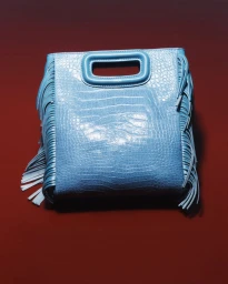 Light blue croc-embossed leather handbag with side fringe detailing placed against a deep red background.