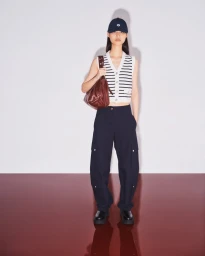Model Jinrong Huang poses in a striped knit vest, navy cargo pants, baseball cap, and burgundy bag against a white wall and maroon floor.