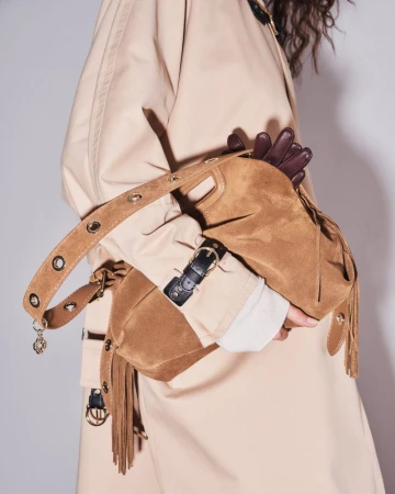 Close-up of a model wearing a beige trench coat while holding a tan suede fringe handbag with eyelet-studded strap details.