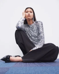 Model seated wearing a silver sequin top and black pants against a white background