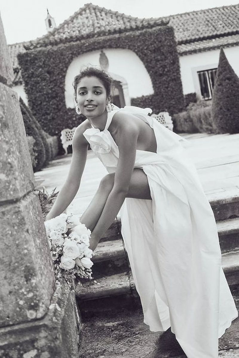 Monochrome outdoor portrait of a woman in an off-white gown, sitting on stone steps with a bouquet, with a building and greenery.