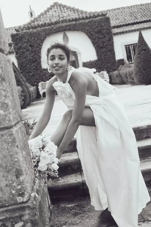 Monochrome outdoor portrait of a woman in an off-white gown, sitting on stone steps with a bouquet, with a building and greenery.