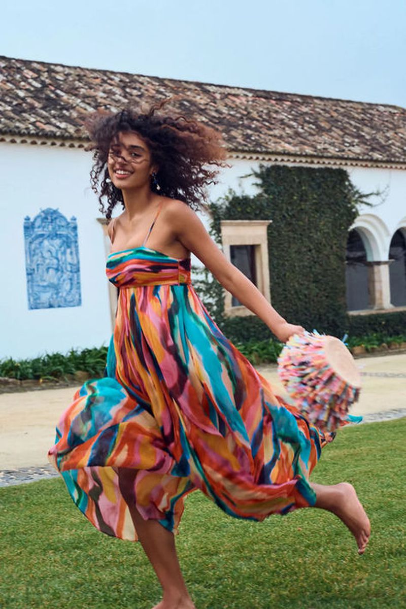 A young woman with curly hair in a colorful dress is running barefoot on a lawn near a building. She is holding a fringed bag.