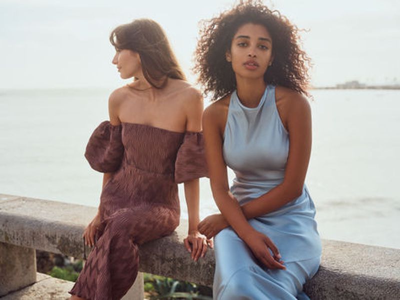 Two women pose on a stone wall near the ocean; one with long brown hair in an off-the-shoulder dress, one with curly hair in a blue dress.