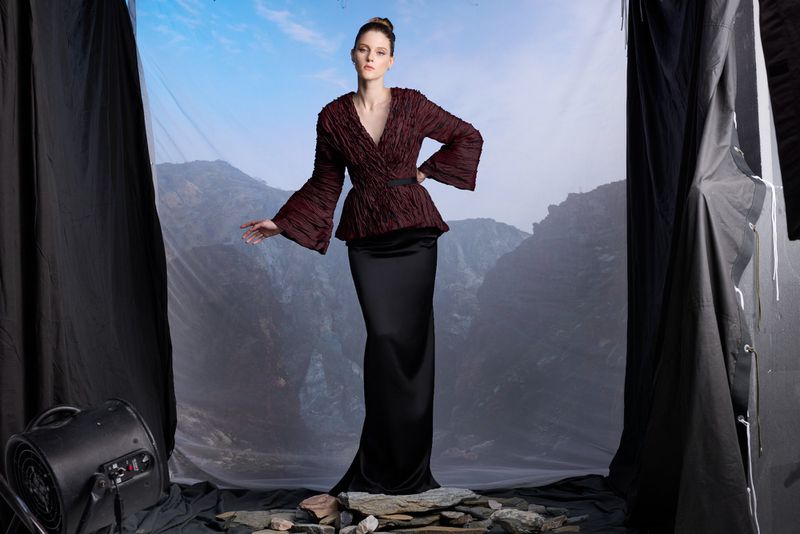 A woman in a burgundy jacket and long black skirt poses in a photo studio with a landscape backdrop and dramatic stage lighting.