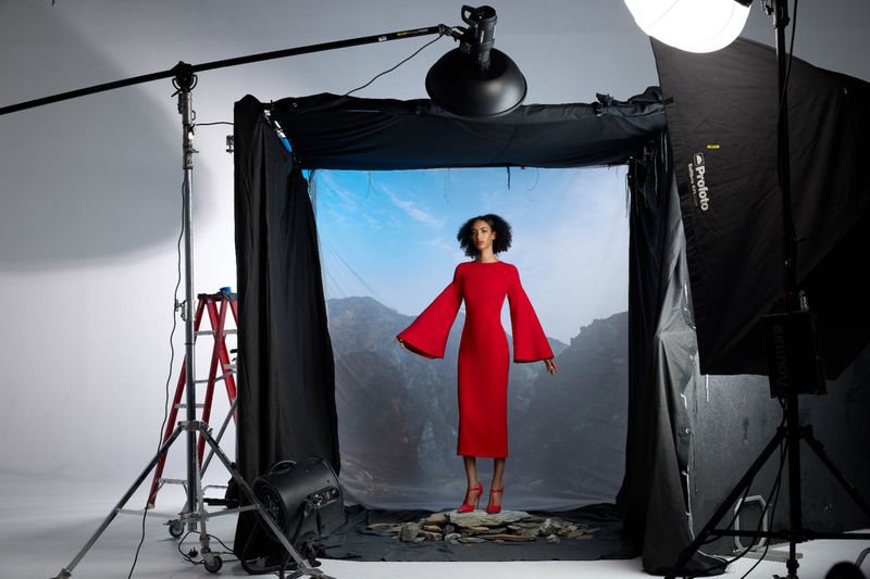 Woman in a red dress poses in a photo studio with lighting equipment, on a rock surface in front of a mountain backdrop.