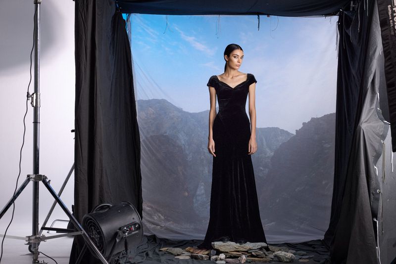Elegant woman in long black velvet gown poses in a photo studio with a scenic mountain backdrop and dramatic black curtains.