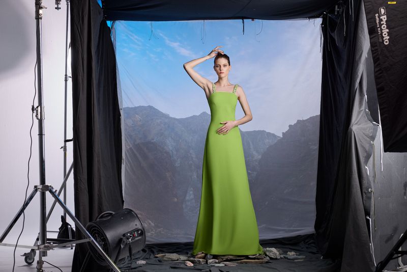 A woman poses in a light green gown in a photo studio with a mountain backdrop and professional lighting equipment visible.