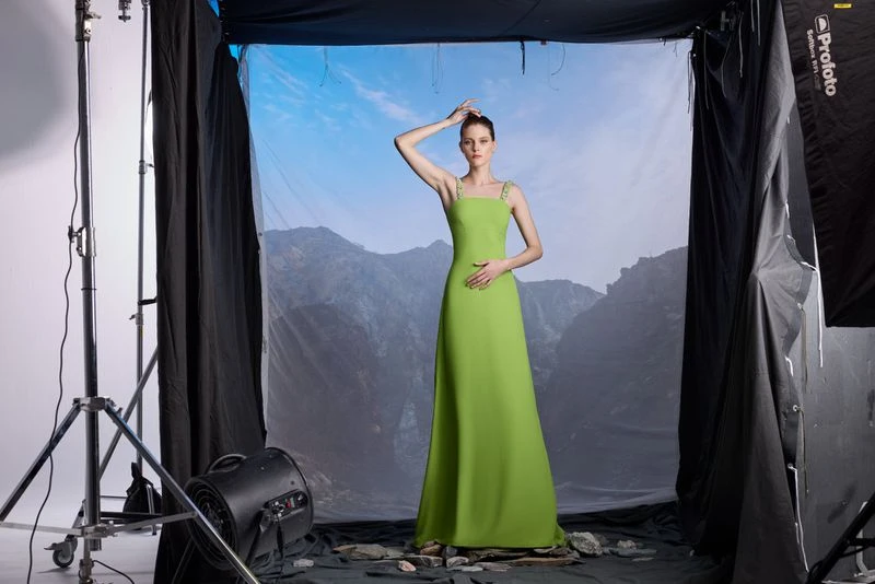 A woman poses in a light green gown in a photo studio with a mountain backdrop and professional lighting equipment visible.