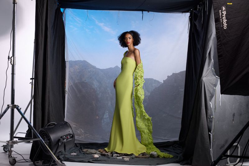 A striking fashion photograph featuring a woman in a green gown posing against a mountain backdrop inside a professional photo studio.
