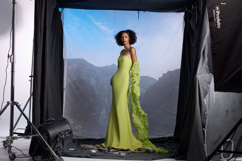 A striking fashion photograph featuring a woman in a green gown posing against a mountain backdrop inside a professional photo studio.