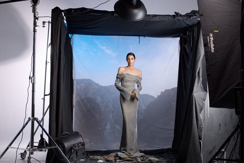 Fashion shot of a woman in a dress against a backdrop of a mountain range, in a photo studio with lighting equipment visible.