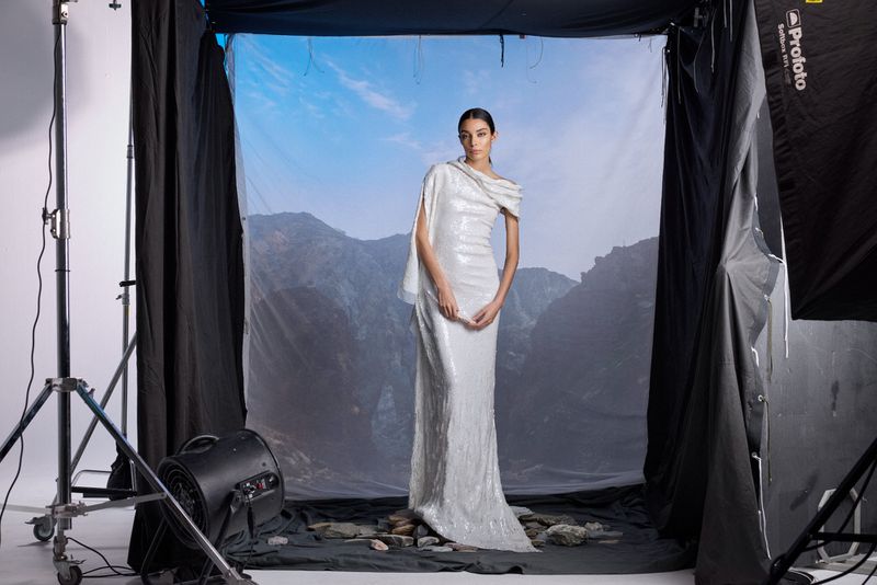 Elegant woman in a sequined gown poses in a studio with a mountain backdrop and professional lighting equipment visible.