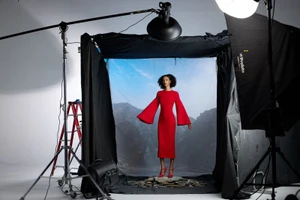 Model Yelibe Tilahun poses in a red bell-sleeve midi dress on a studio set with scenic mountain backdrop and photography lighting.