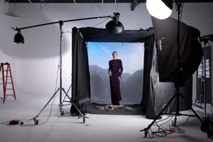 Model Yelibe Tilahun poses in a long plum gown inside a studio setup surrounded by lighting gear and a mountain backdrop.
