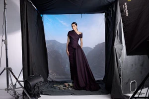 Model Yelibe Tilahun wearing a draped deep purple off-the-shoulder gown standing inside a studio photo set with landscape backdrop.