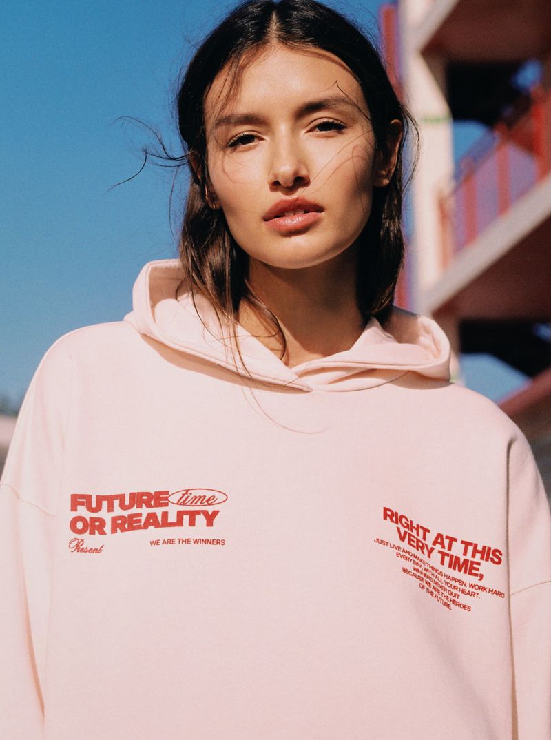 Portrait of a brunette fashion model in a pink hoodie with graphic text, photographed outdoors with a clear blue sky.