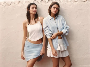 Two models in light blue and white summer outfits posing against a textured off-white wall.