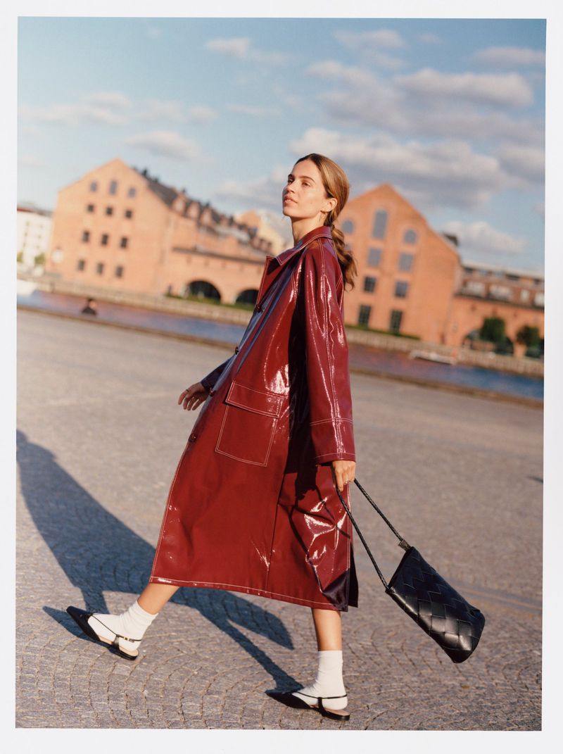 A stylish woman walks across a cobblestone square with brick buildings under a blue sky, wearing a red coat and carrying a black purse.