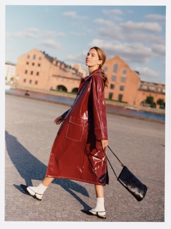 A stylish woman walks across a cobblestone square with brick buildings under a blue sky, wearing a red coat and carrying a black purse.