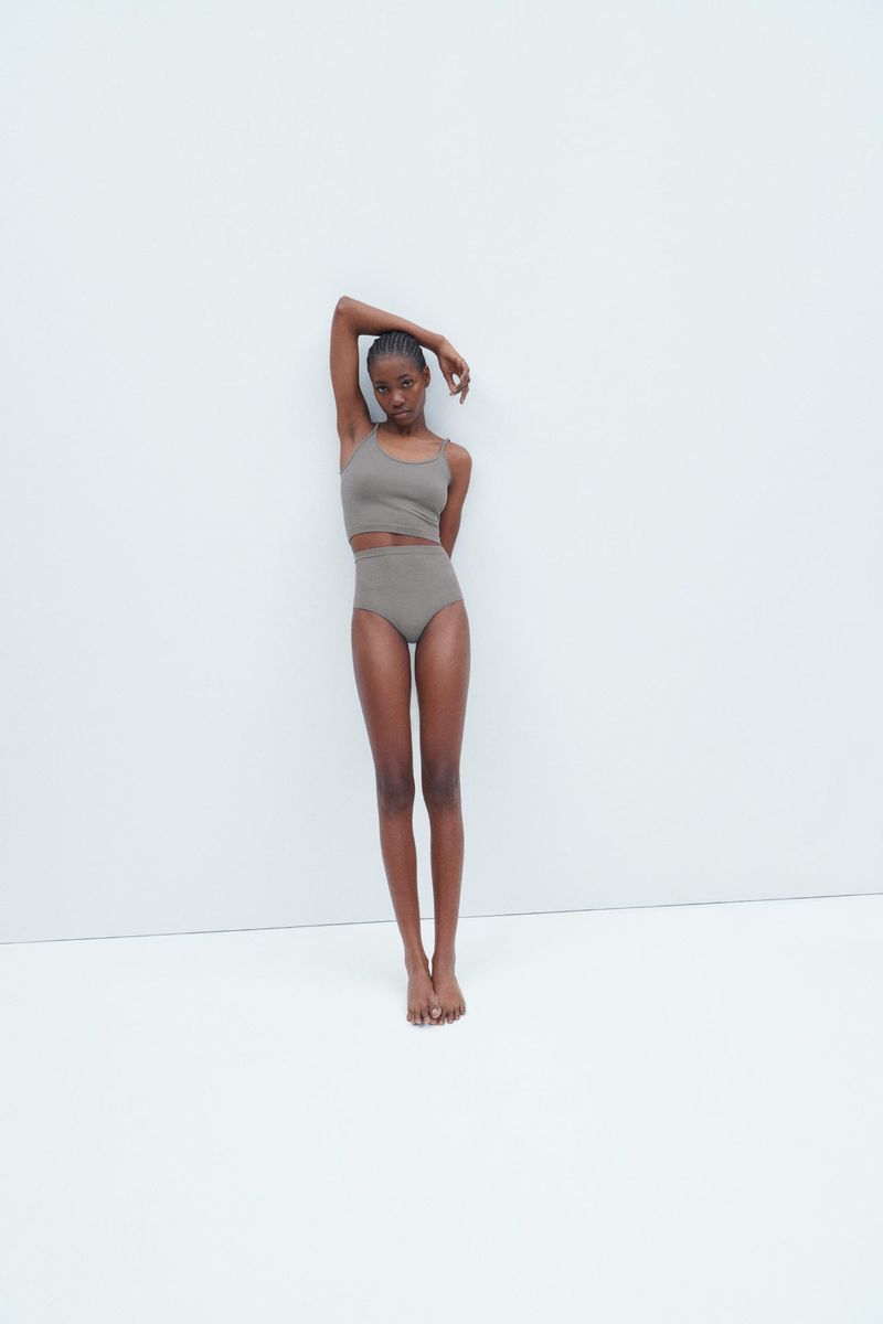 A slender, dark-skinned woman in a taupe-colored crop top and high-waisted briefs poses against a stark white wall in a fashion forward portrait.