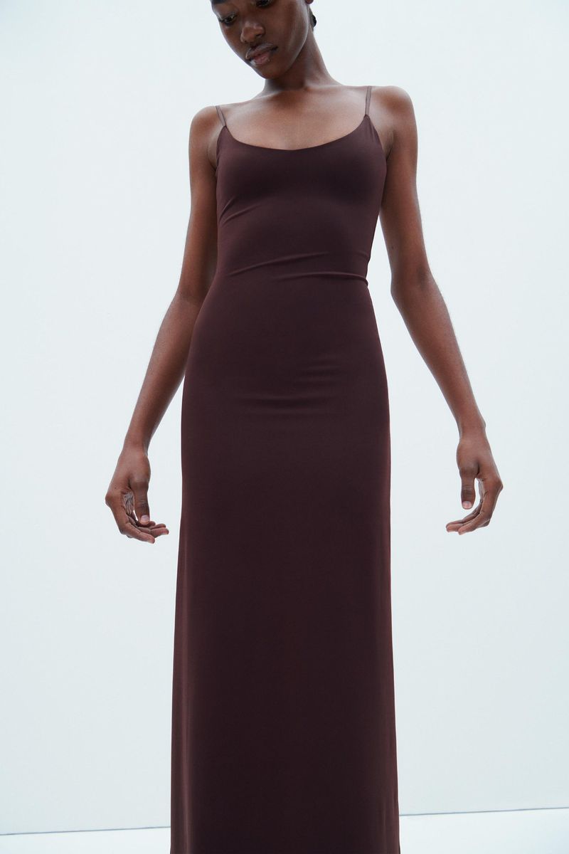 Woman in a brown, floor-length spaghetti strap dress posing against a white background. She has dark skin and her arms are outstretched.