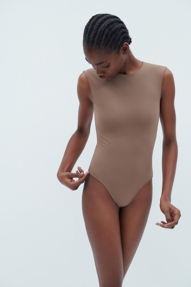 A slender African-descent female fashion model poses in a neutral taupe bodysuit against a stark white background in a full body shot.