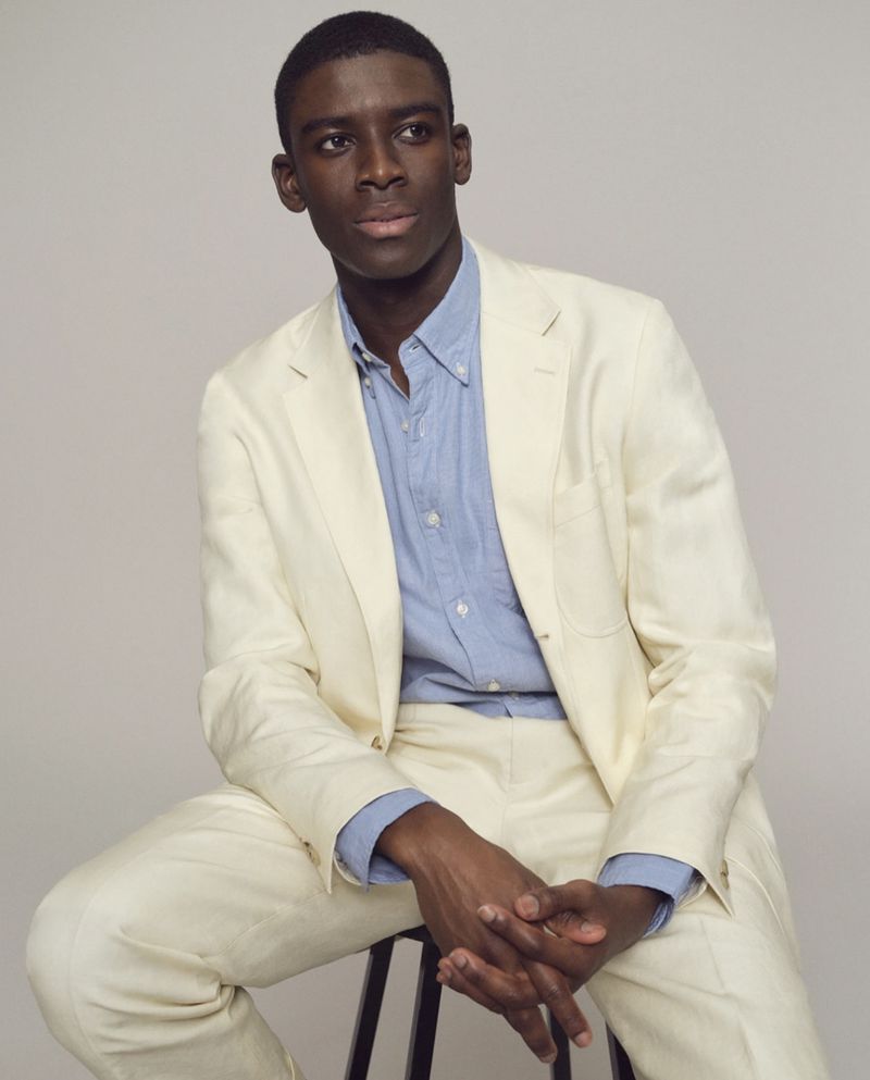 Model Karl-Yann Assah seated on a stool wearing a cream tailored suit and light blue button-down shirt against a neutral studio backdrop.