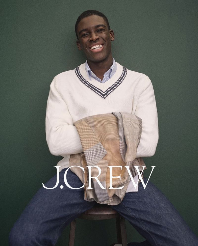 Smiling Black man poses in J. Crew V-neck sweater, blue jeans, holding jacket while seated on a stool against a dark green background.