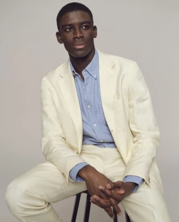Model Karl-Yann Assah seated on a stool wearing a cream tailored suit and light blue button-down shirt against a neutral studio backdrop.