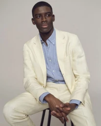 Model Karl-Yann Assah seated on a stool wearing a cream tailored suit and light blue button-down shirt against a neutral studio backdrop.