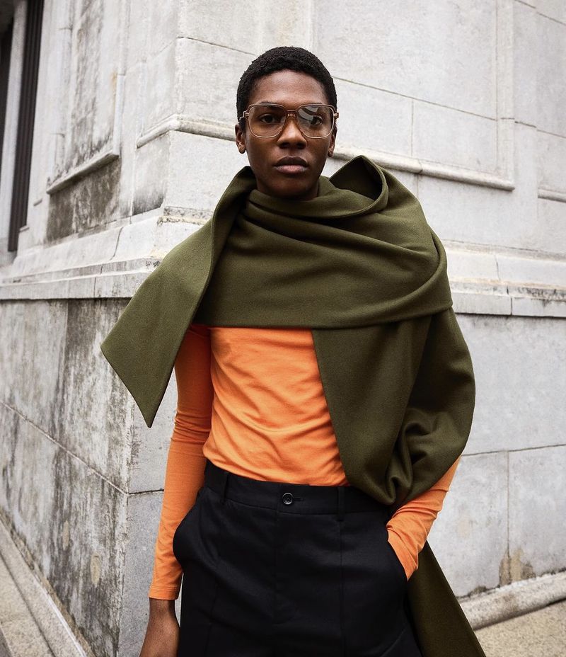 Man in orange shirt and black pants draped with a green scarf stands against a light stone building in an urban environment.