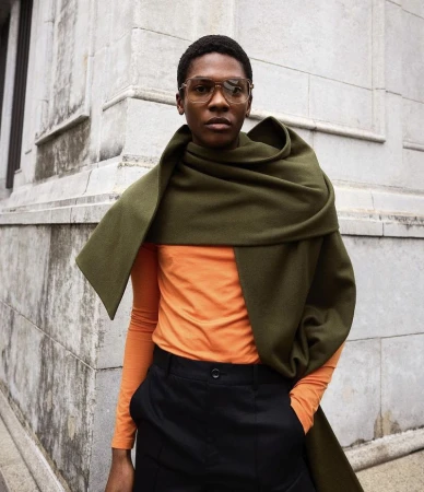 Man in orange shirt and black pants draped with a green scarf stands against a light stone building in an urban environment.