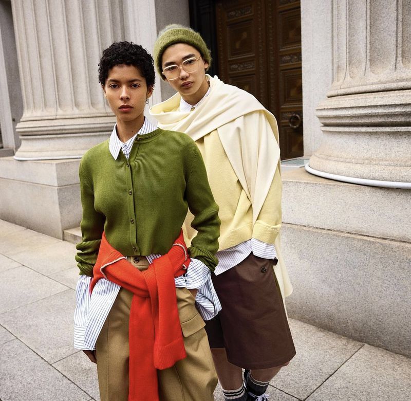 Two stylish people pose outdoors, modeling olive green cardigan, orange sweater, striped button-down shirt, and khaki shorts for a fashion shoot.