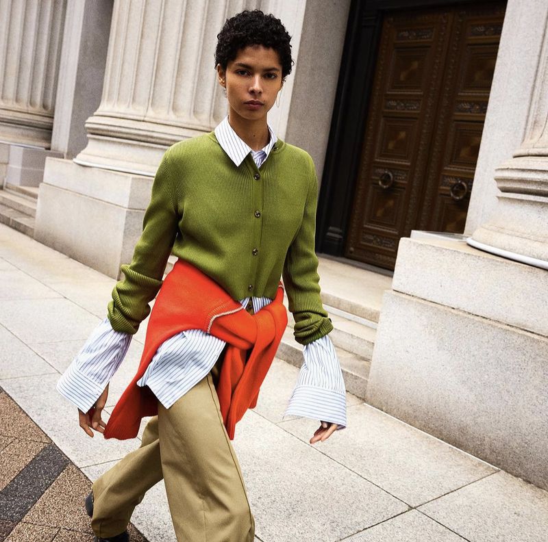 Fashion model in an olive cardigan, layered shirts, and beige trousers walking on a city sidewalk near building columns and doors.