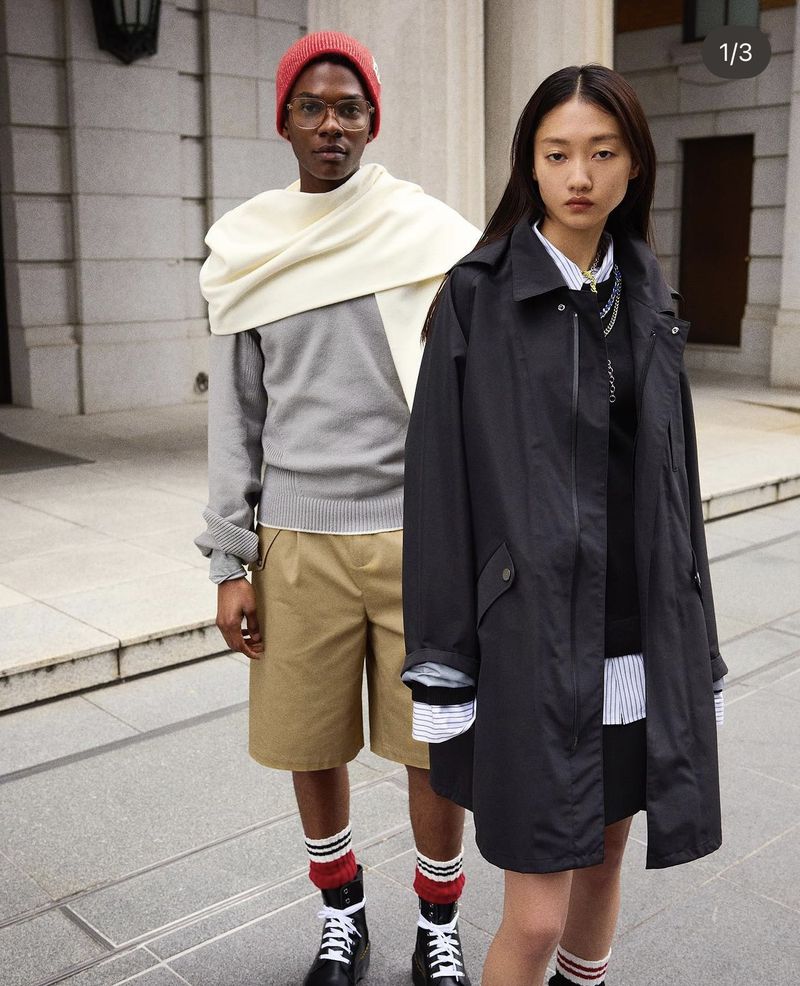 Two fashion models posing outdoors in jackets, sweaters, shorts, and boots, standing on a concrete surface with a building backdrop.