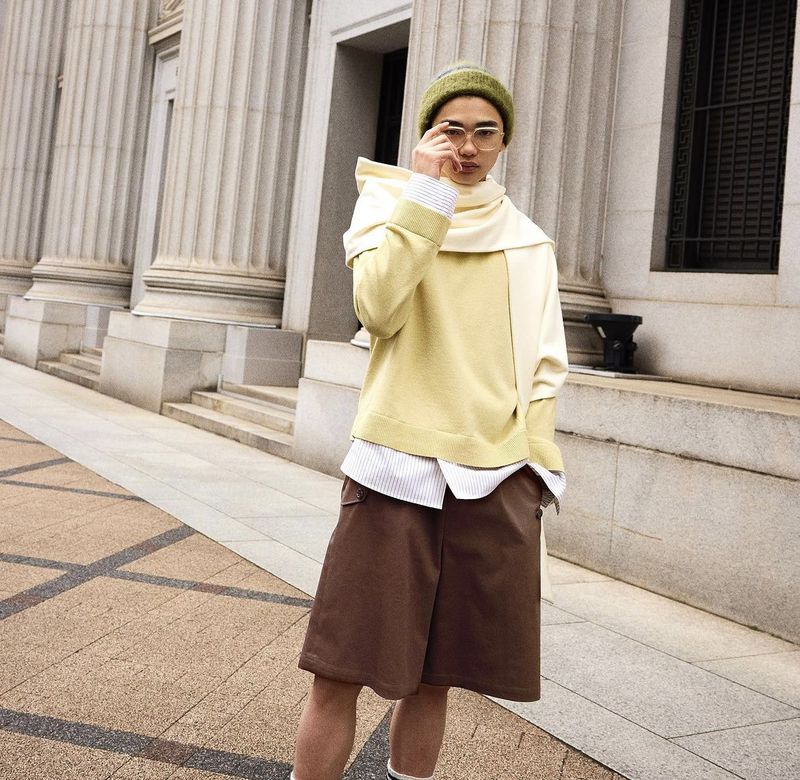 Man in streetwear poses outdoors in a yellow sweater, brown shorts, olive beanie, and eyeglasses, with classical building background.