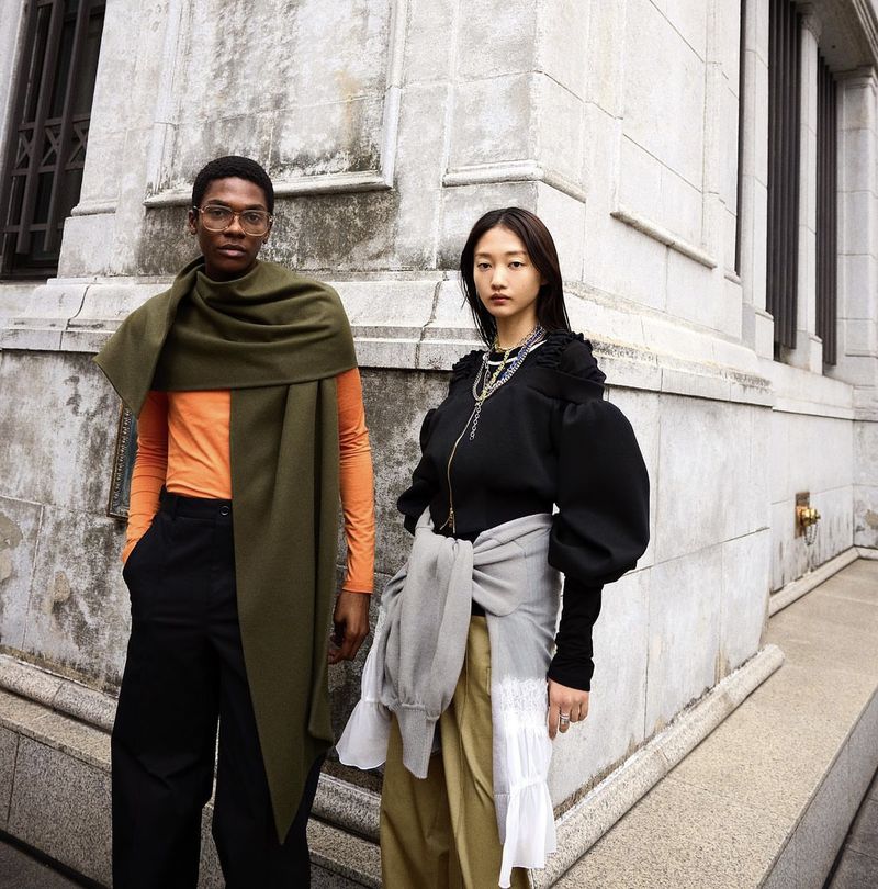 Two fashion models pose outdoors wearing layered autumn knitwear and oversized draped garments against a stone building exterior.